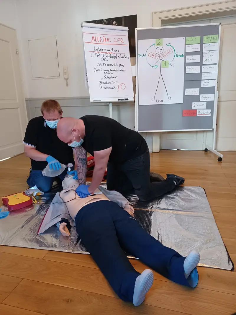 Zwei Helfer mit Mundschutz versorgen ein Übungsphantom mit Beatmungsbeutel und Defibrillator