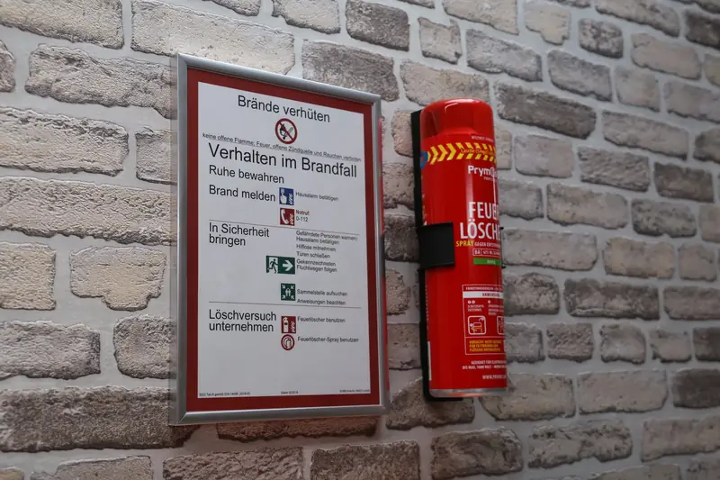 Prymos-Feuerlöschspray in der Wandhalterung neben dem Aushang Verhalten im Brandfall